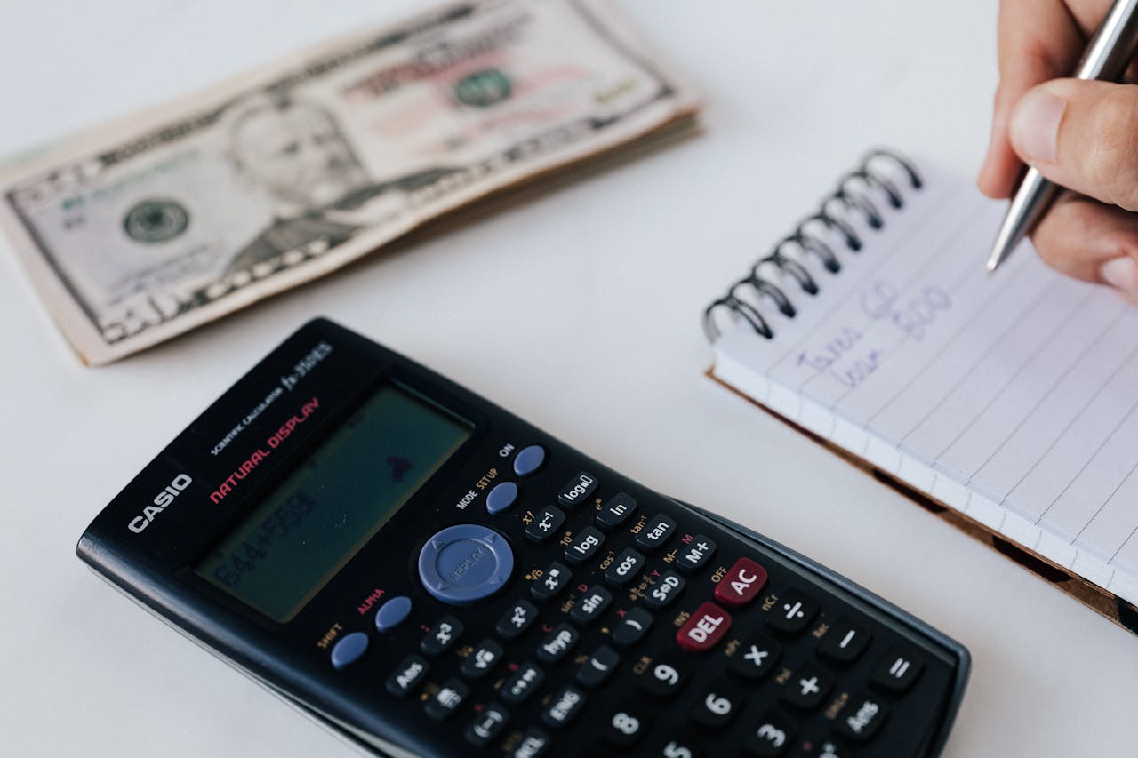 Close-up of a calculator, US dollar bill, and a hand writing in a notebook, symbolizing financial planning.