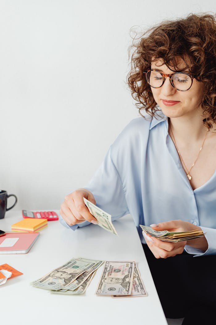 Caucasian woman in blue top counting money at desk with eyeglasses and a necklace.